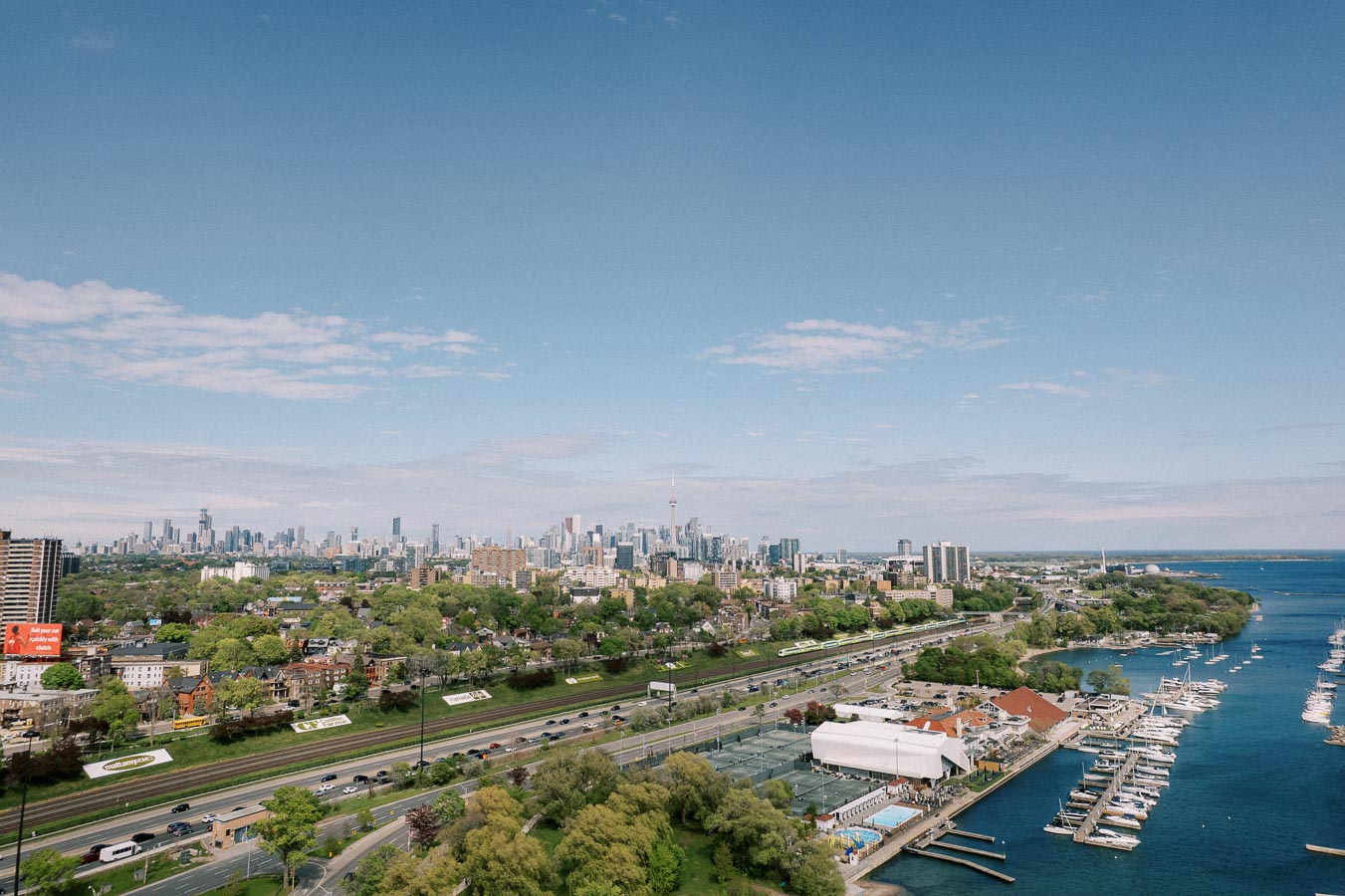 Aerial view of a cityscape with a prominent skyline, waterfront, and marina on a clear day, featuring green spaces and buildings, set against a blue sky with some clouds.