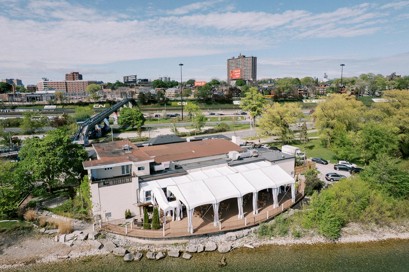 Aerial view of a waterfront restaurant with a white canopy-covered outdoor seating area, surrounded by lush greenery and overlooking a calm body of water. In the background, urban buildings and a highway are visible under a partly cloudy sky.