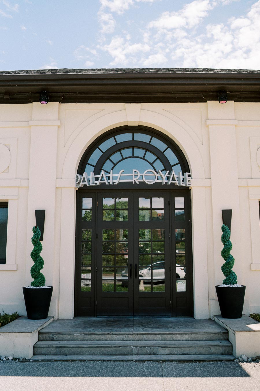 Front entrance of Palais Royale with arched glass door and topiary plants, under a clear blue sky.