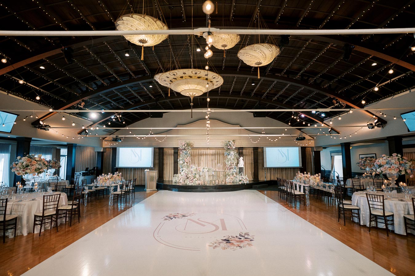 Elegant wedding reception hall with a beautifully decorated stage, floral centerpieces, string lights, and a polished dance floor with monogram.