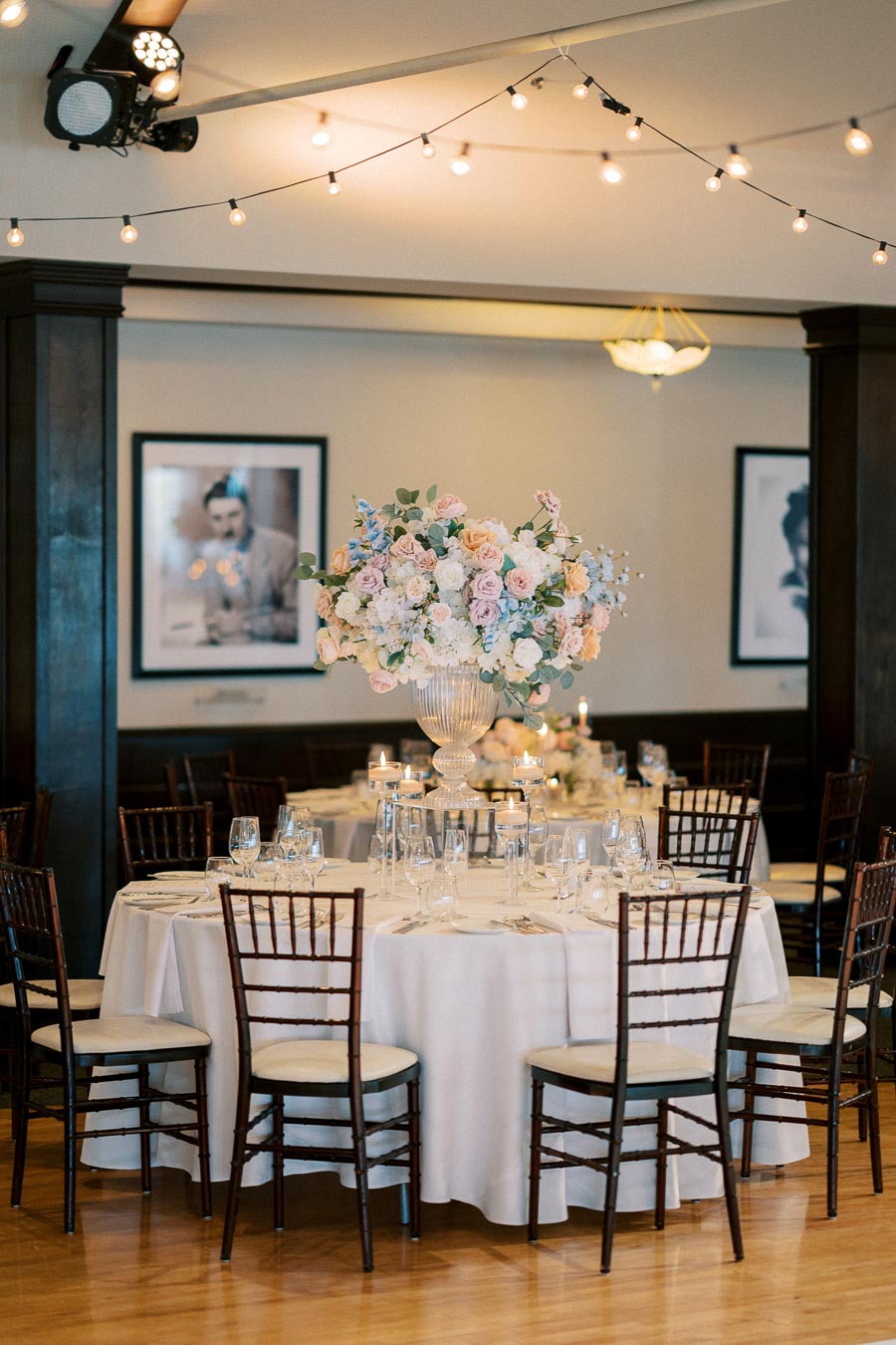 Elegant wedding reception table with a large floral centerpiece, surrounded by wooden chairs. The table is set with glassware, plates, and cutlery under ambient string lights in a stylish venue.