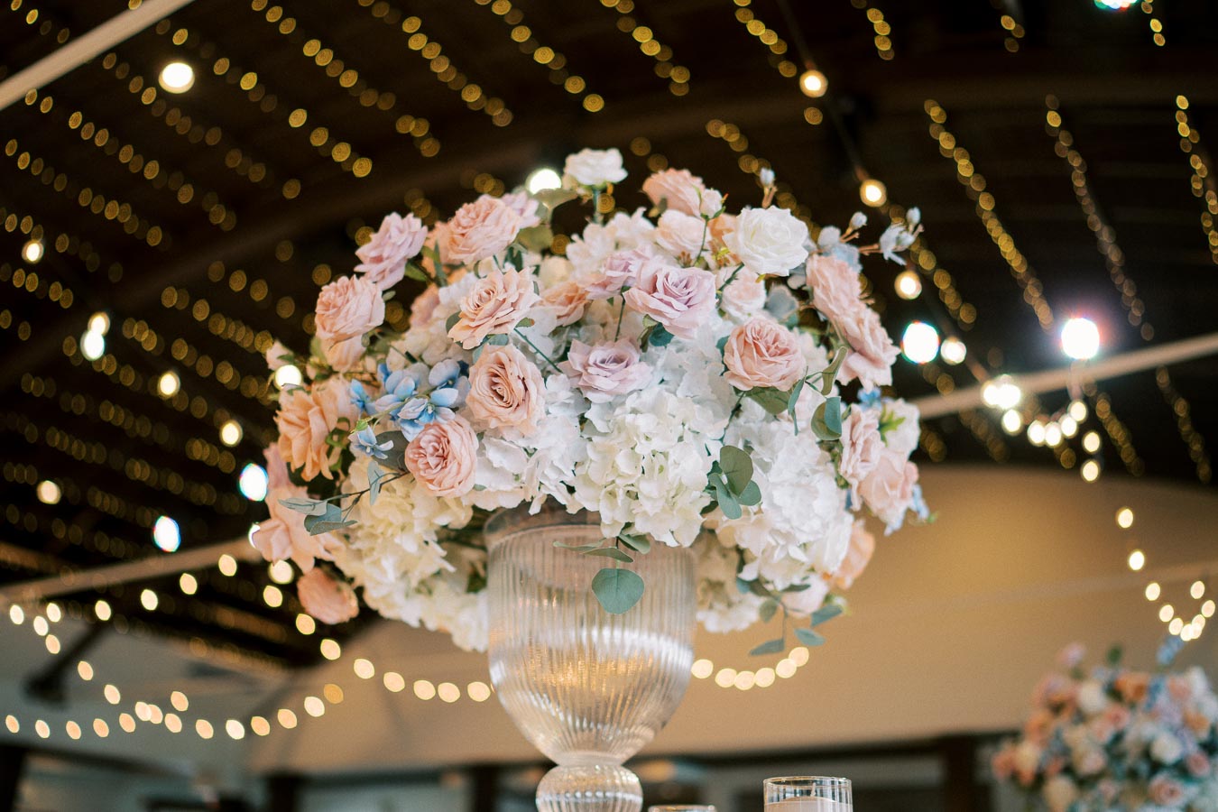 Elegant wedding centerpiece featuring a tall glass vase with a lush arrangement of pale pink roses, white hydrangeas, and greenery, set against a warmly lit venue with twinkling string lights.