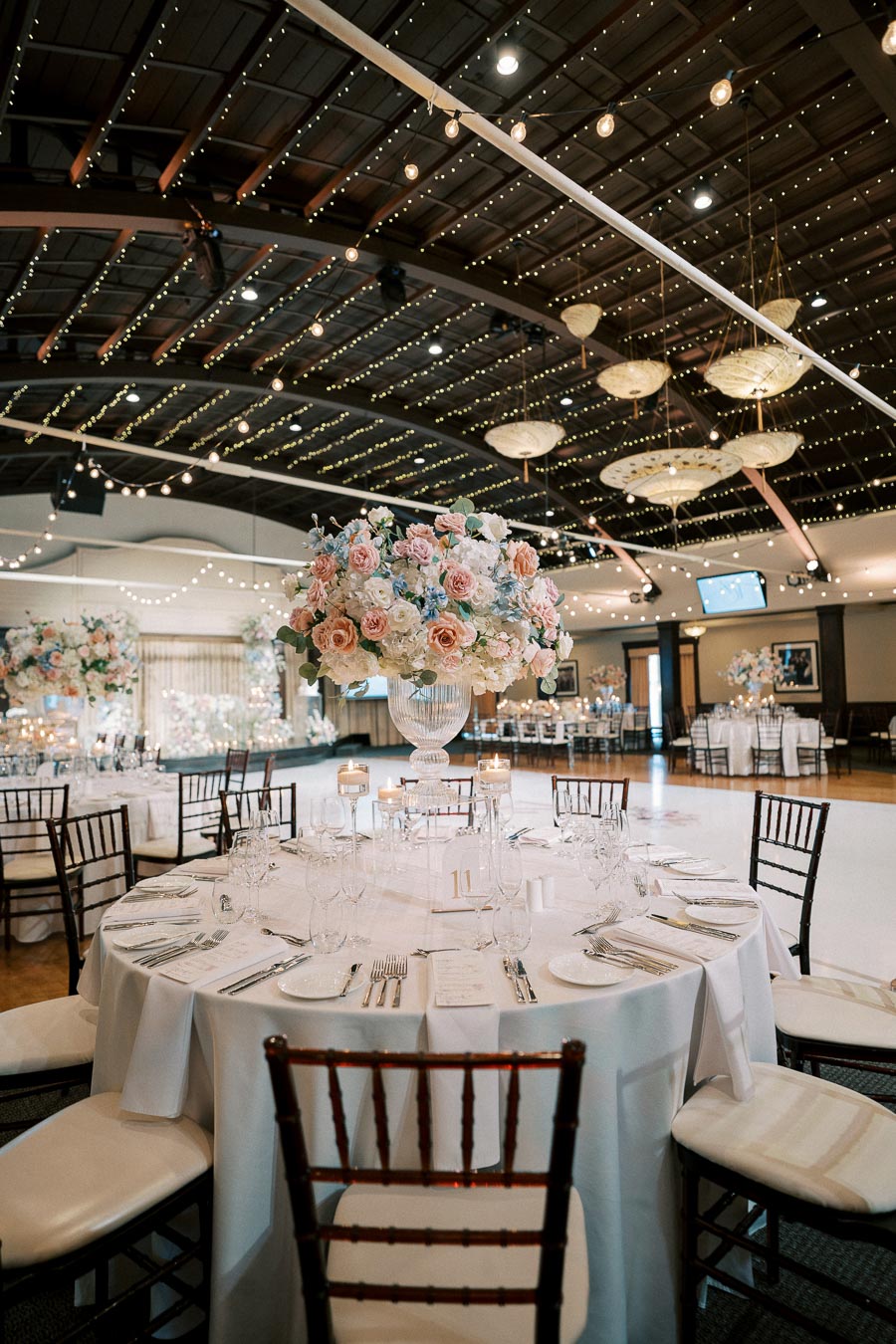 Luxurious wedding reception venue with elegant table settings, featuring a centerpiece of pastel-colored flowers and surrounded by wooden chairs. The ceiling is adorned with ambient string lights, creating a romantic atmosphere.