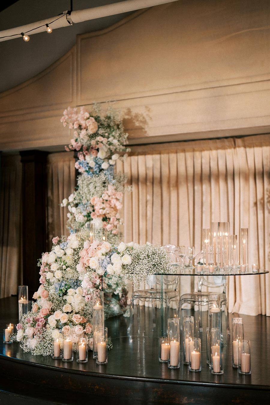 Elegant wedding reception setup featuring a luxurious floral arrangement with pink, white, and blue roses and baby's breath cascading over a glass table. Tall glass candleholders with lit candles adorn the black wooden floor, creating a romantic ambiance. Cream-colored drapery in the background adds to the sophisticated atmosphere.