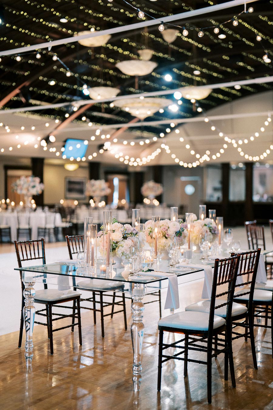 Elegant wedding reception venue with decorative string lights, glass tables, floral centerpieces, and chiavari chairs, creating a sophisticated and romantic ambiance.