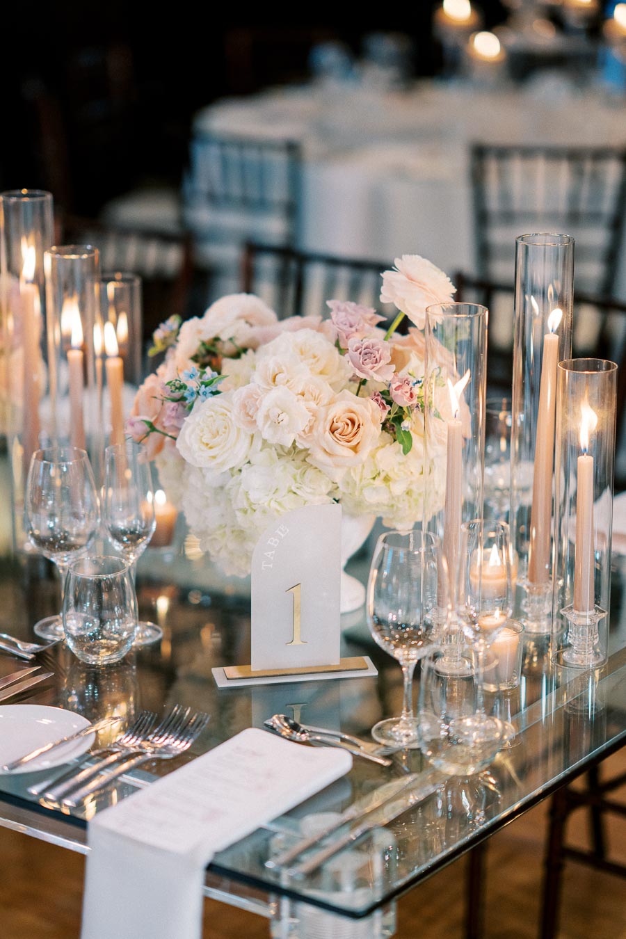 Elegant wedding table setting featuring a centerpiece of white and blush flowers, surrounded by tall candles in glass holders, silverware, and a table number card labeled 1 on a reflective glass table.