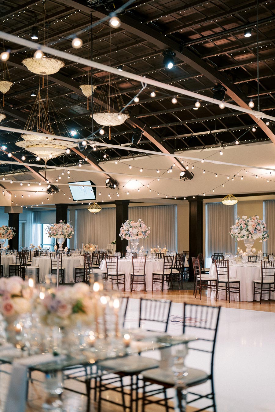 Elegant wedding reception venue with round tables, white tablecloths, and tall floral centerpieces, under a canopy of string lights and decorative chandeliers.