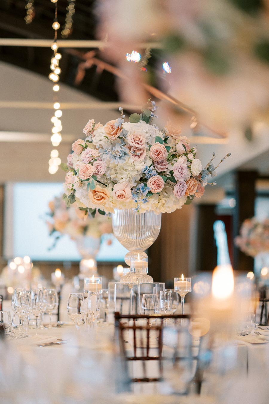 Elegant wedding reception table setting with a large floral centerpiece featuring pink and white roses, surrounded by glassware and lit candles.