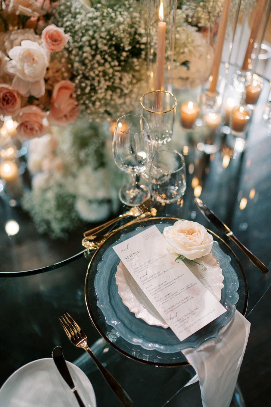 Elegant wedding table setting featuring a floral centerpiece, gold-trimmed glassware, a handwritten menu, and a white rose place card on a blue ceramic plate, illuminated by candlelight for a romantic ambiance.
