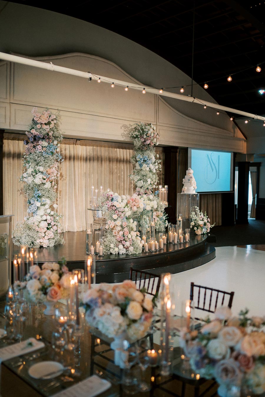 Elegant wedding venue with floral arrangements, featuring tall flower pillars and candles on a dimly lit stage, creating a romantic atmosphere.