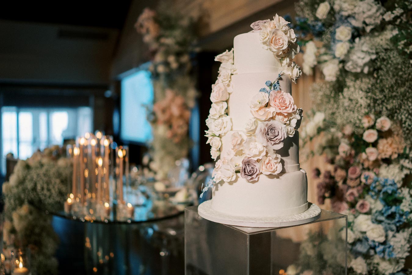 Elegant four-tier white wedding cake adorned with pastel-colored roses and floral decorations, set in a luxurious reception hall with soft candlelight and lush greenery in the background.