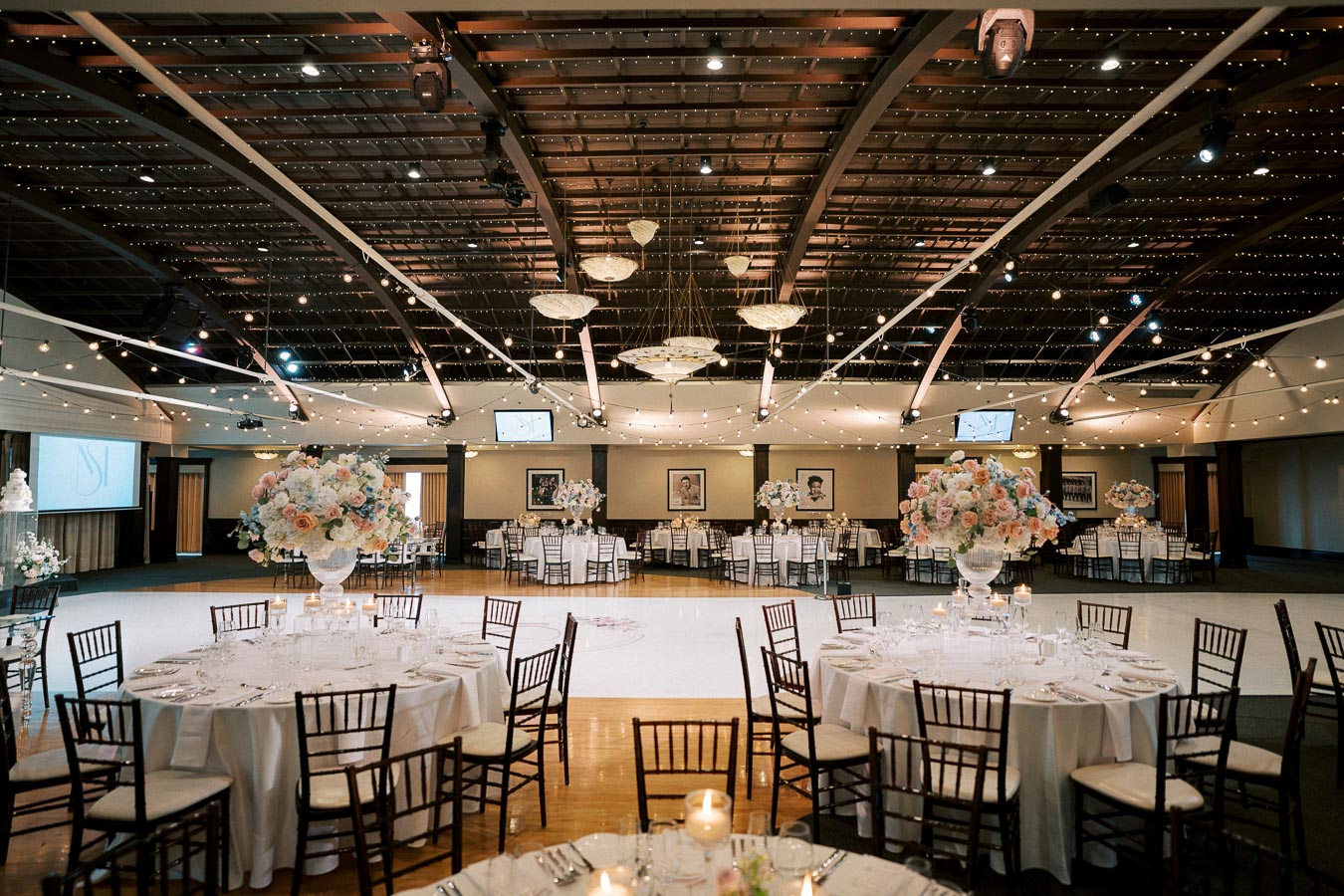 Elegant wedding reception hall with round tables, floral centerpieces, and string lighting under an arched ceiling.