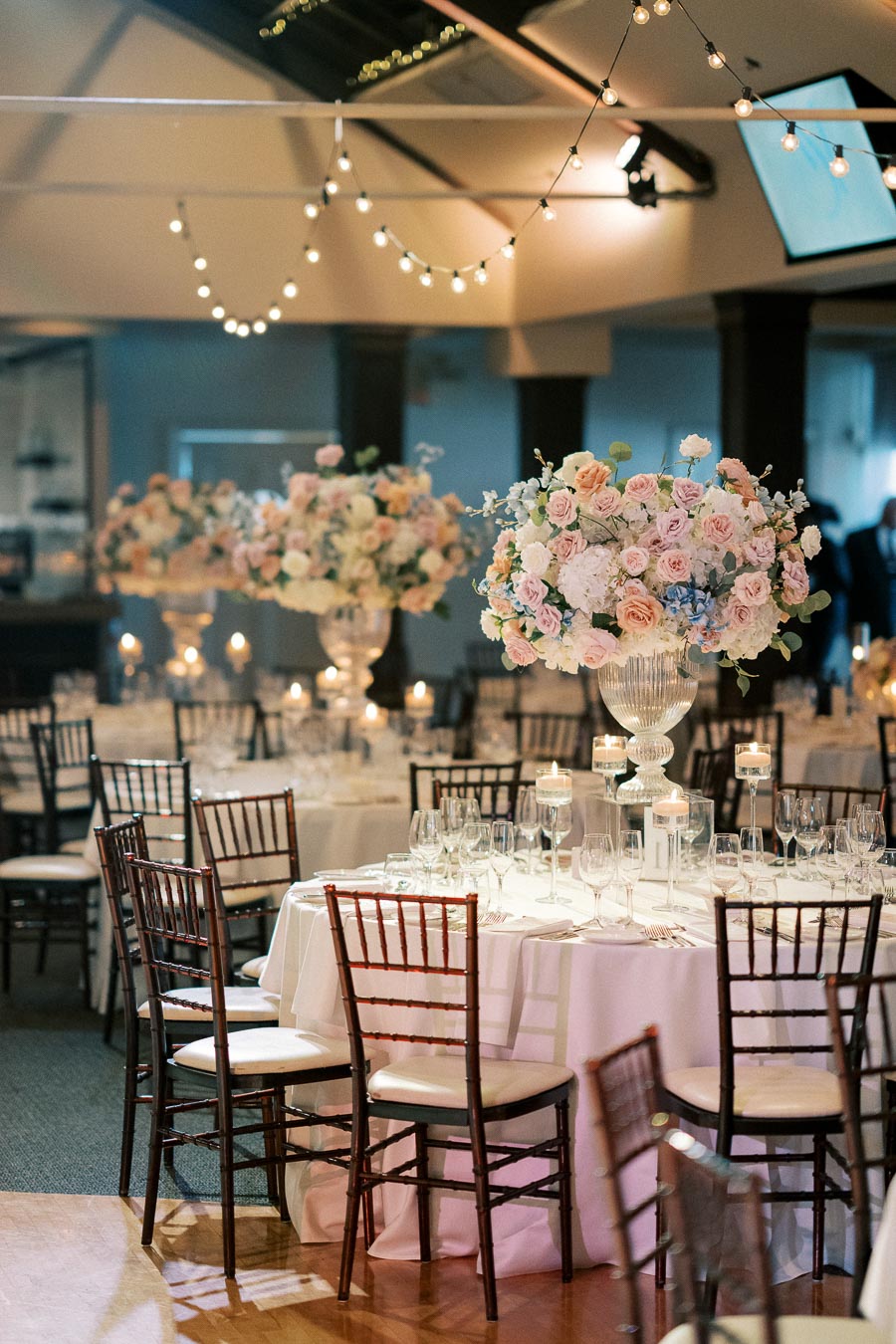 Elegant wedding reception setup featuring round tables adorned with white tablecloths, tall floral centerpieces of pastel roses, and candles, surrounded by wooden chairs under warm string lights.