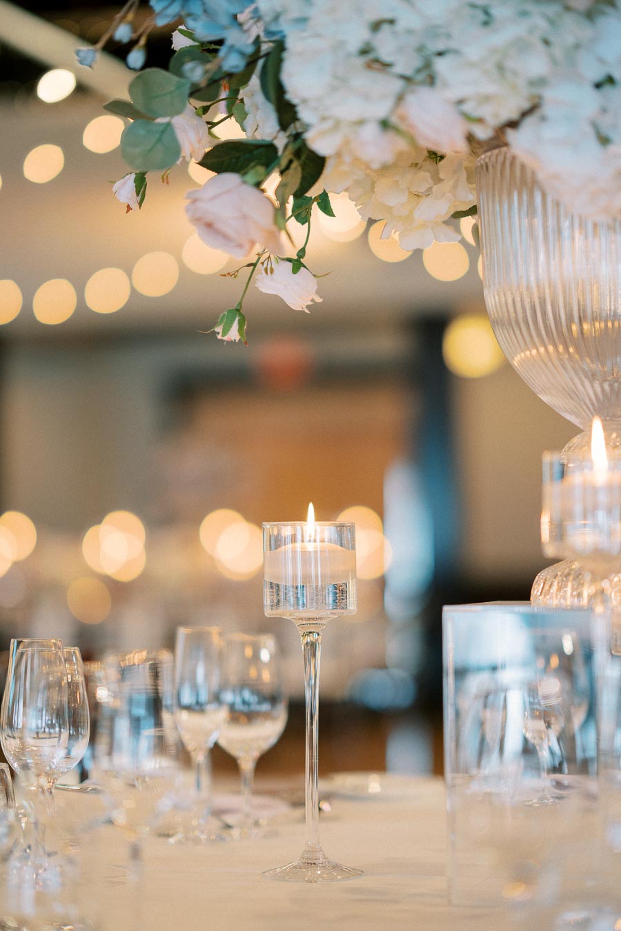 Elegant wedding table setting with a focus on a tall glass candle holder surrounded by wine glasses and a luxurious floral centerpiece, featuring white roses and hydrangeas. Soft bokeh background enhances the romantic ambiance.