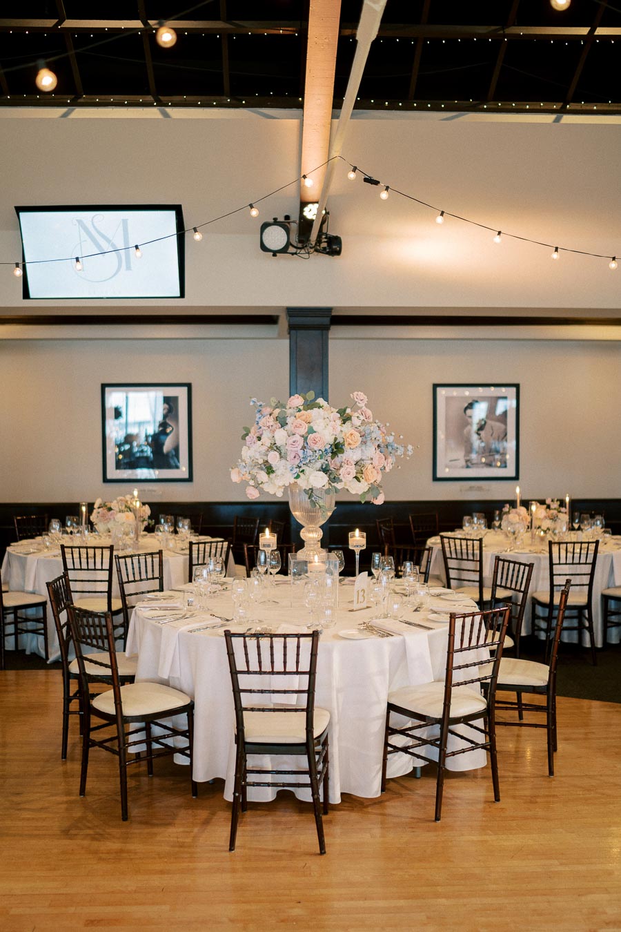 Elegant wedding reception setup with round tables adorned with white tablecloths, floral centerpieces, and candle holders, surrounded by wooden chairs under ambient string lighting.