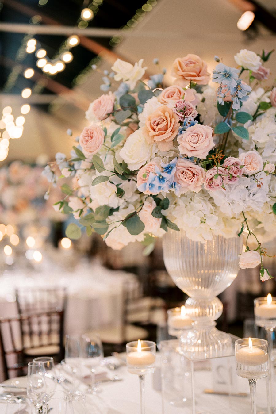 Elegant wedding table centerpiece with a bouquet of pink and white roses, blue flowers, and greenery in a tall vase, surrounded by lit candles.
