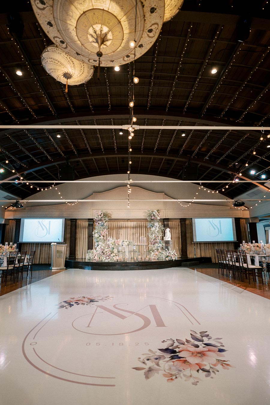 Elegant wedding reception venue featuring a beautifully decorated dance floor with floral designs and initials, stunning floral arrangements on stage, and ornate hanging light fixtures.