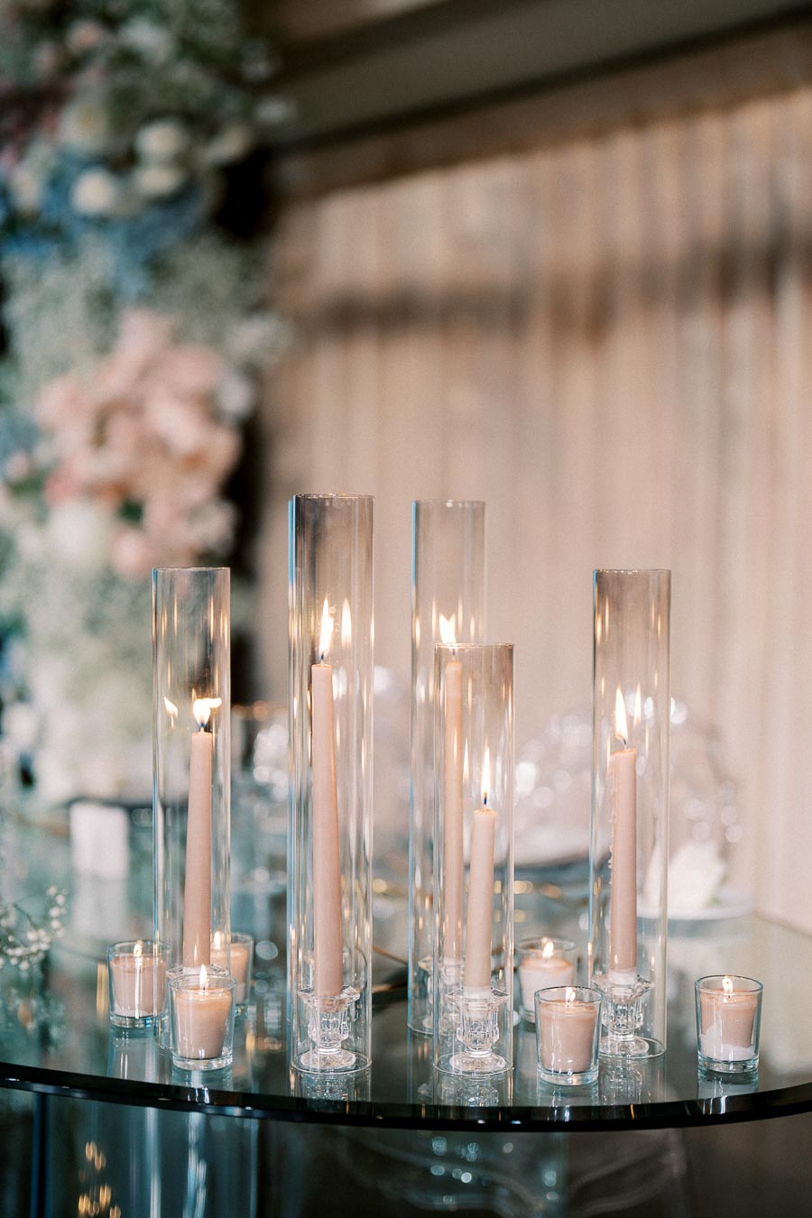 Elegant glass cylinders with lit candles on a glass table, surrounded by soft-focus floral décor, creating a romantic and serene atmosphere.
