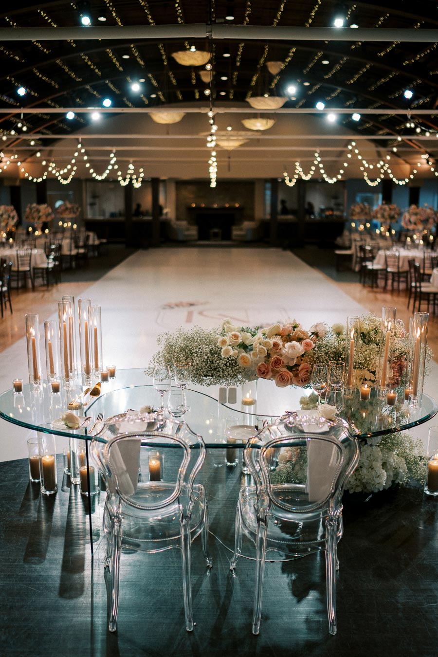 Elegant wedding reception venue with a beautifully set sweetheart table, adorned with floral arrangements and candlelight, overlooking an expansive, decorated dance floor and ambient string lights.