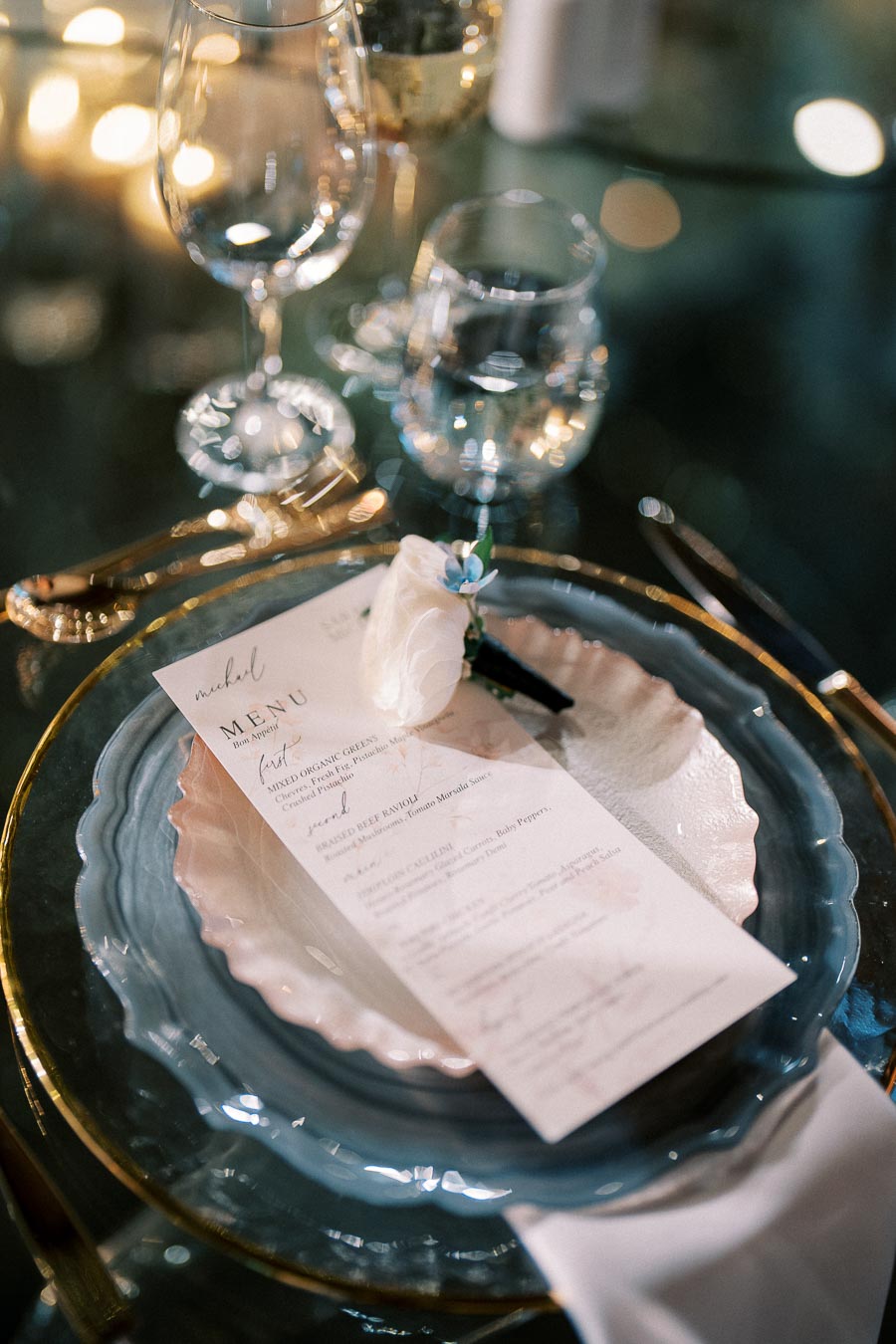 Elegant dinner table setting with a decorative menu card on a white and blue plate, accompanied by gold silverware and a white rose accent; wine glass and water glass in the background, illuminated by soft ambient lighting.