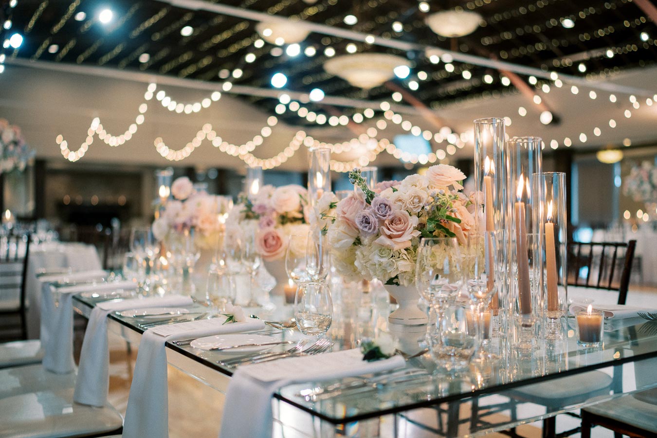 Elegant wedding reception setup featuring a long table adorned with pastel floral arrangements, candles in glass holders, and string lights creating a romantic ambiance in the background.
