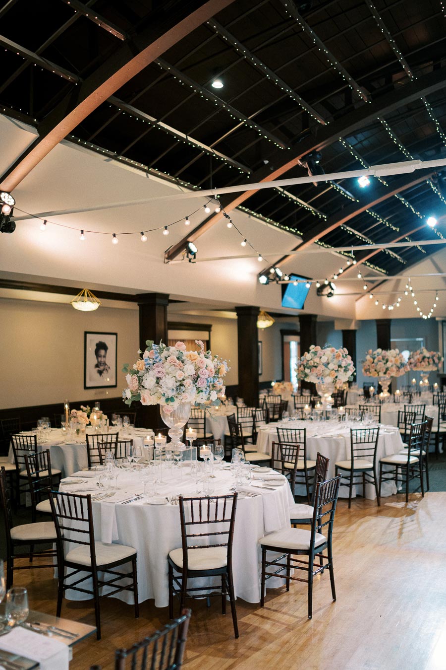 Elegant wedding reception hall with round tables draped in white linens, decorated with tall floral centerpieces and surrounded by wooden chairs. Warm string lights hang from the high ceiling, creating a romantic ambiance.