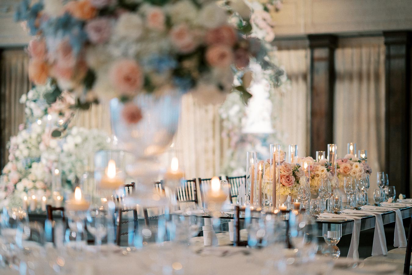 Elegant wedding reception table decorated with white and pastel flowers, surrounded by lit candles and glassware, creating a romantic atmosphere.