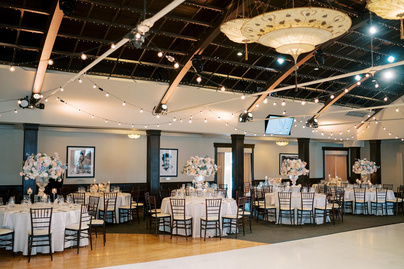 Elegant banquet hall with round tables set for a wedding reception, adorned with white tablecloths and floral centerpieces, under a ceiling of string lights.