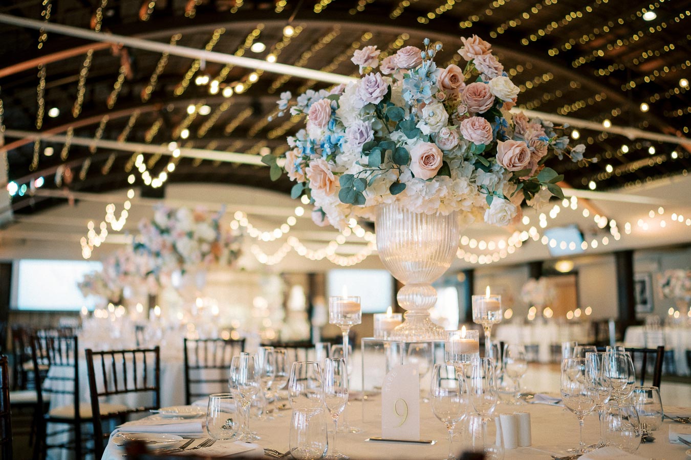 Elegant wedding reception with a beautifully decorated table centerpiece featuring pastel roses and greenery in a glass vase, surrounded by sparkling wine glasses, candles, and warm string lights creating an inviting ambiance.