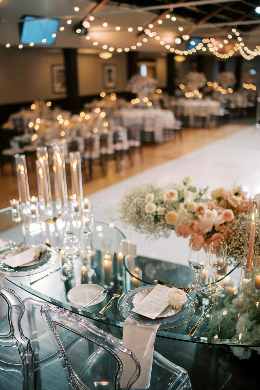 Elegant wedding reception setting featuring a beautifully decorated table with clear glass plates, elegant glassware, white napkins, and a centerpiece of delicate peach and white flowers. Surrounding the area are soft, glowing candle lights and romantic string lights, creating a warm and inviting atmosphere for a celebration.