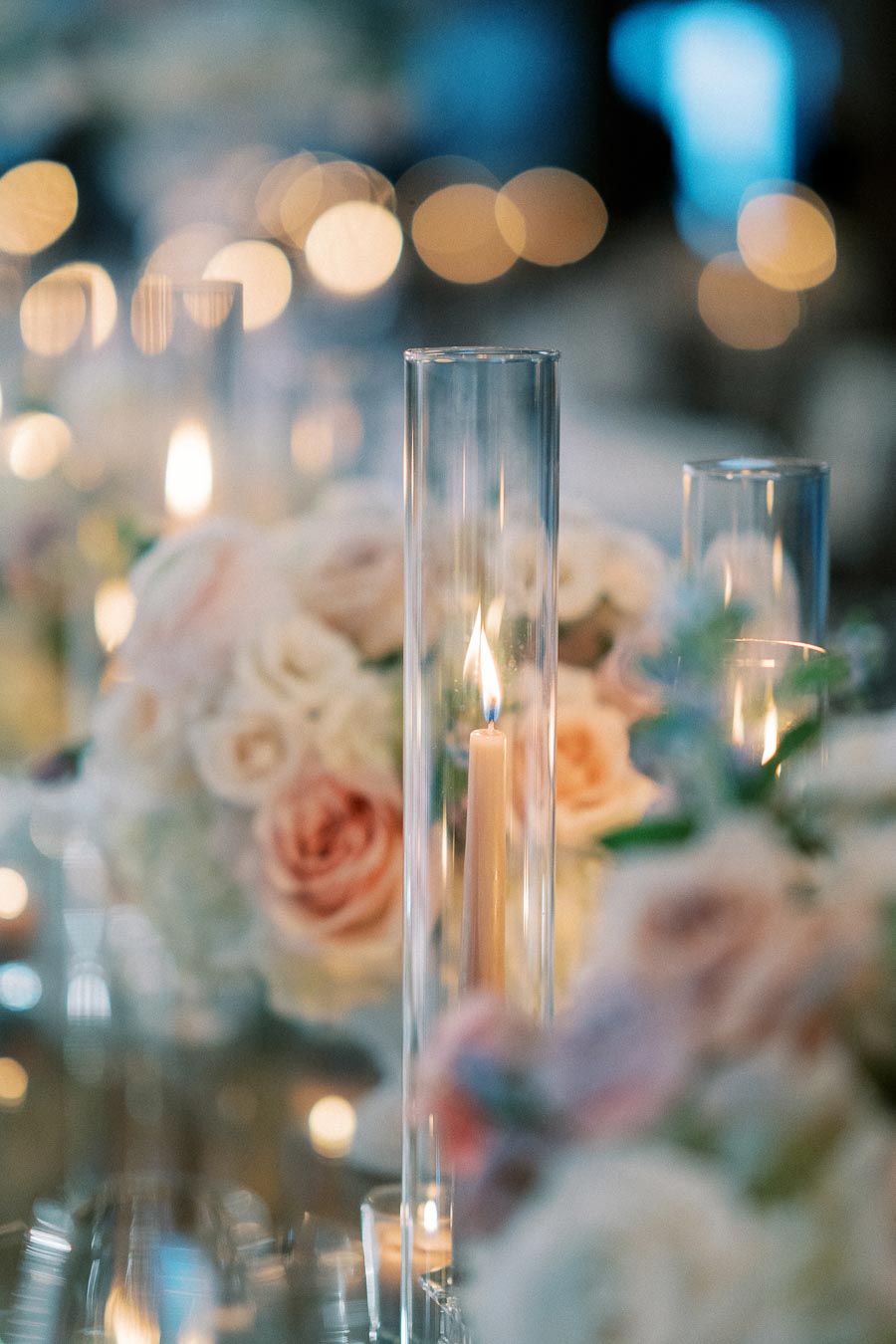 Elegant wedding table centerpiece featuring tall, lit candles in glass holders surrounded by soft, pink and white floral arrangements, creating a romantic and luxurious ambiance.