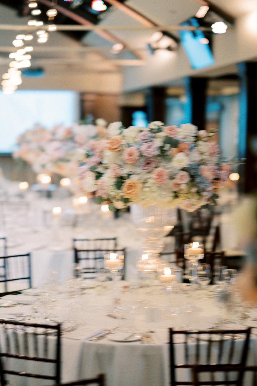 Blurred image of an elegant wedding reception setup featuring a round table with a white tablecloth, decorated with tall floral centerpieces of pastel roses and hydrangeas, surrounded by candles in glass holders, creating a romantic and sophisticated ambiance.