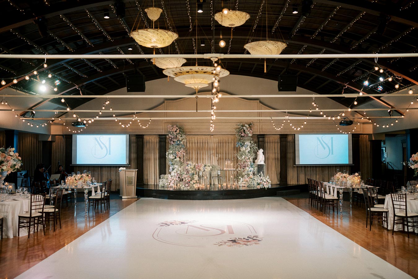 Luxurious wedding reception hall with elegant floral decorations and romantic string lights, featuring beautifully set tables and a polished dance floor, creating a sophisticated and enchanting atmosphere.