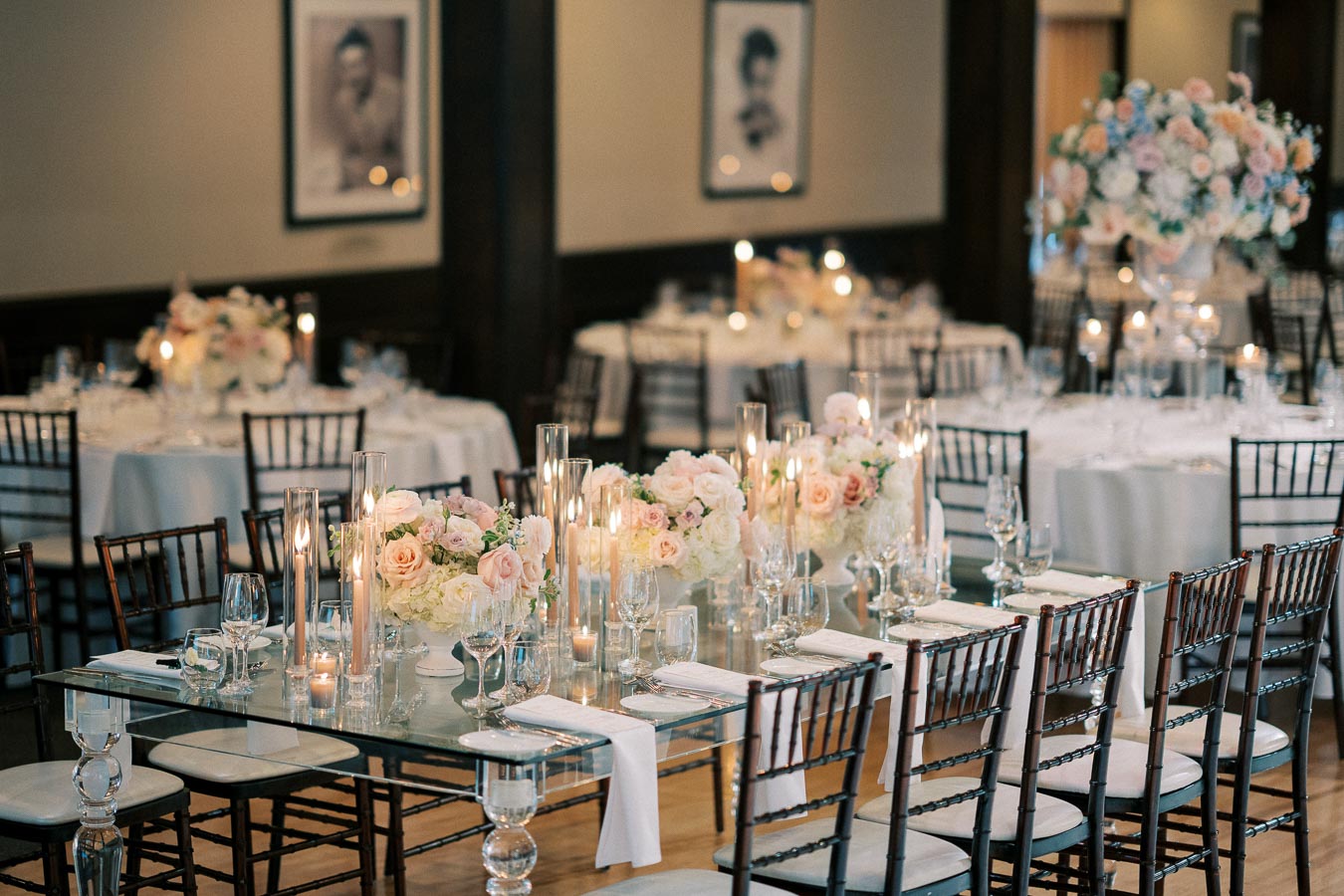 Elegant wedding reception setup featuring a glass dining table with floral centerpieces of pink and white roses, surrounded by dark wooden chairs, in a softly lit venue with framed photographs on the walls.