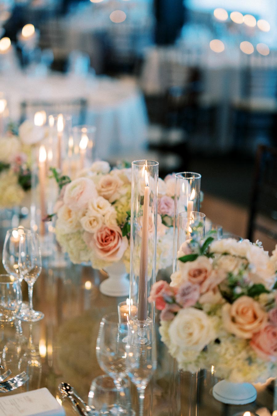 Elegant wedding table setting with soft pink and white floral arrangements, surrounded by tall, thin candles, creating a romantic ambiance in a dimly lit reception hall.
