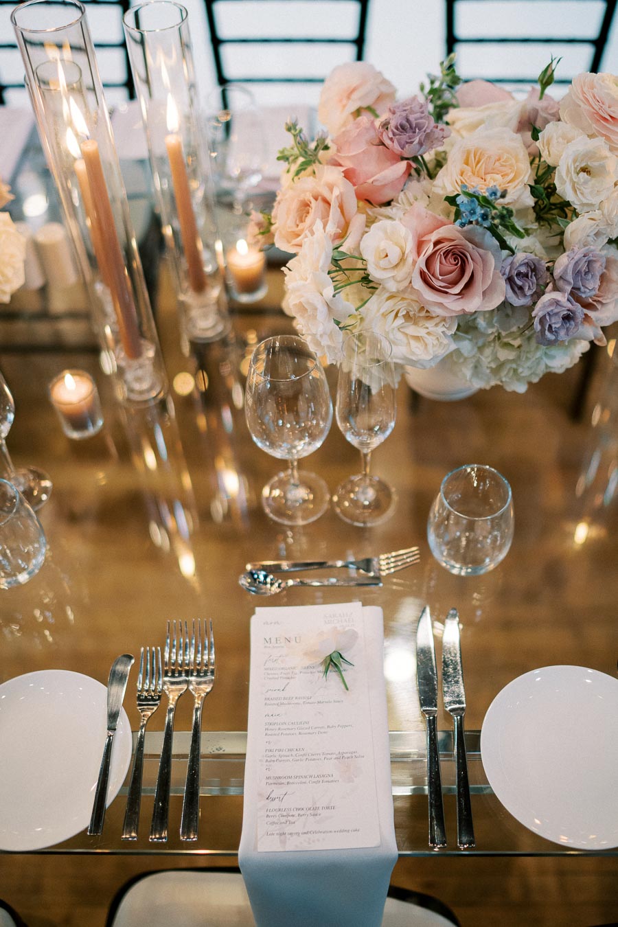Elegant wedding table setting with a floral centerpiece of pastel roses and candles, featuring silver cutlery, wine glasses, and a neatly arranged menu on a white tablecloth.
