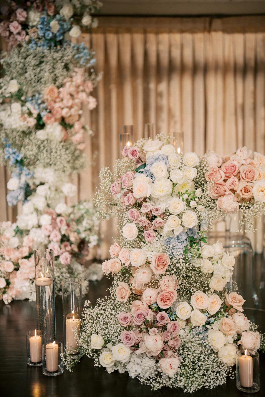 Elegant wedding floral arrangement featuring pastel roses and baby's breath with tall candles in glass holders on a dark wooden table.