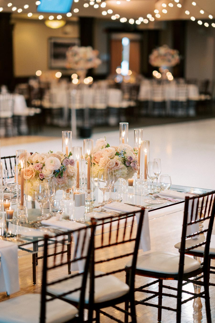 Elegant wedding reception table setting with floral arrangements, tall candles, and ambient string lights in a beautifully decorated venue.