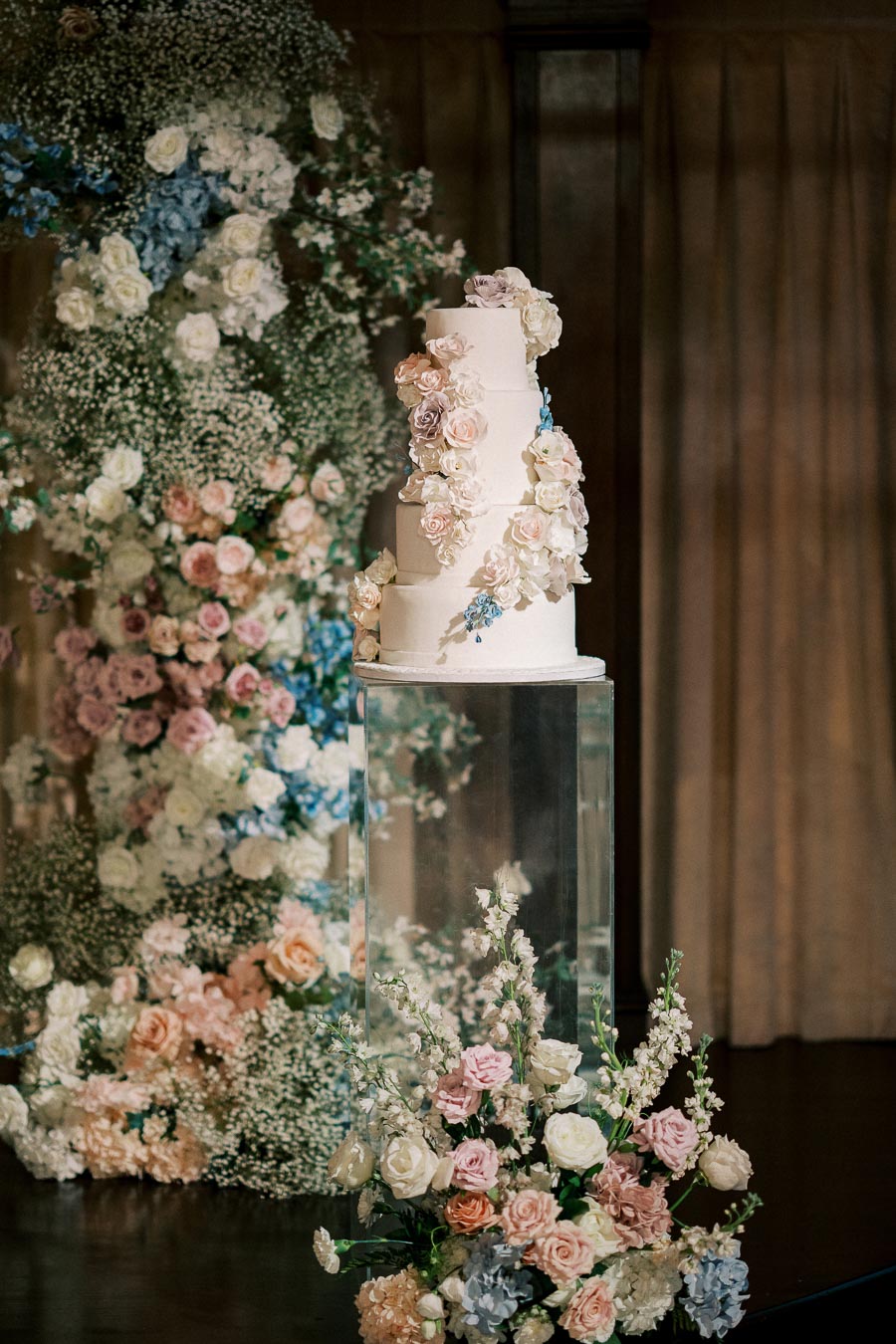 A three-tiered wedding cake adorned with pastel-colored floral arrangements, displayed on a glass pedestal, surrounded by an elegant backdrop of lush flowers, creating a romantic and sophisticated setting.