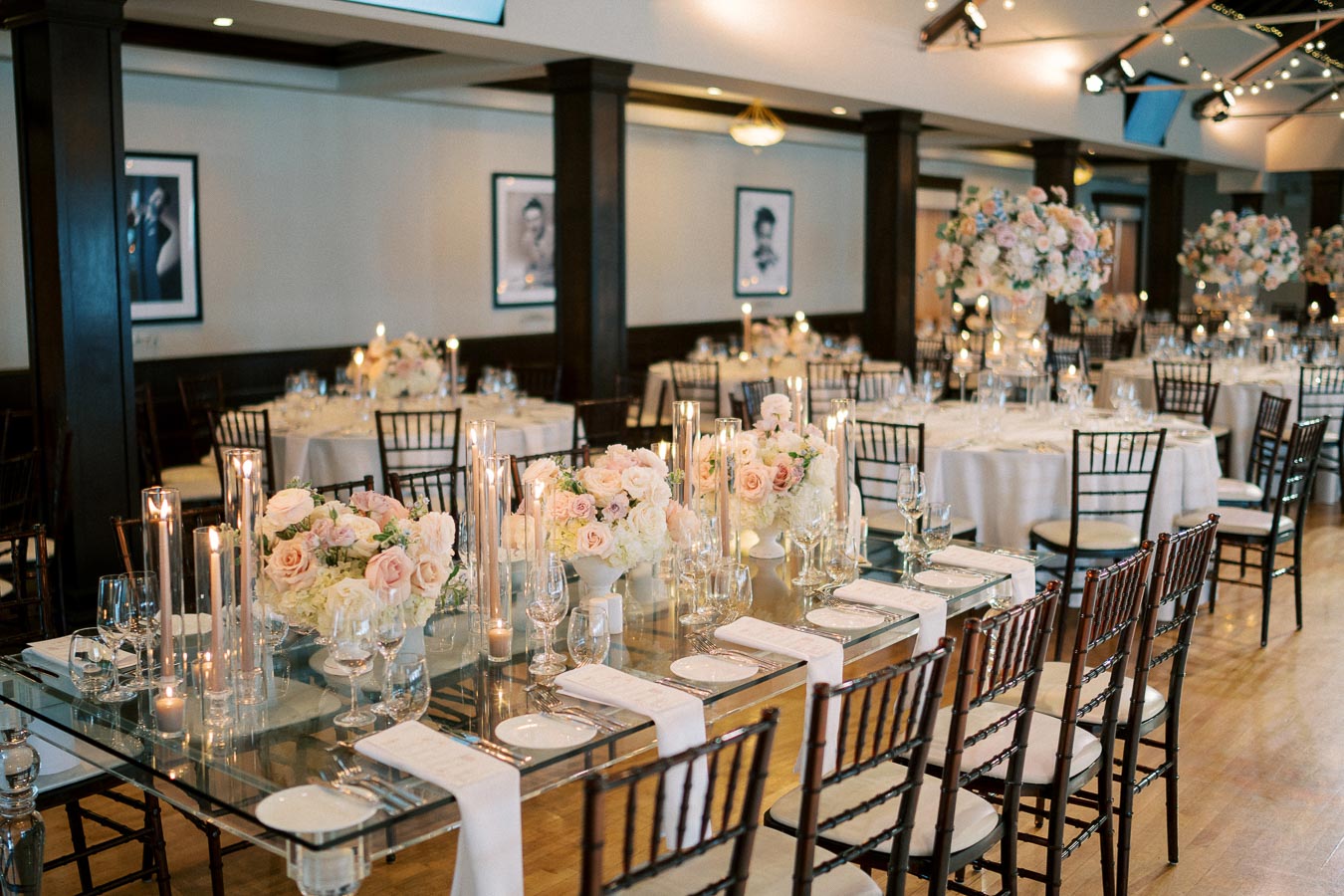 Elegant wedding reception venue with beautifully decorated tables featuring floral arrangements, candles, and neatly arranged place settings, complemented by chic wooden chairs and ambient lighting, creating a sophisticated and romantic atmosphere.
