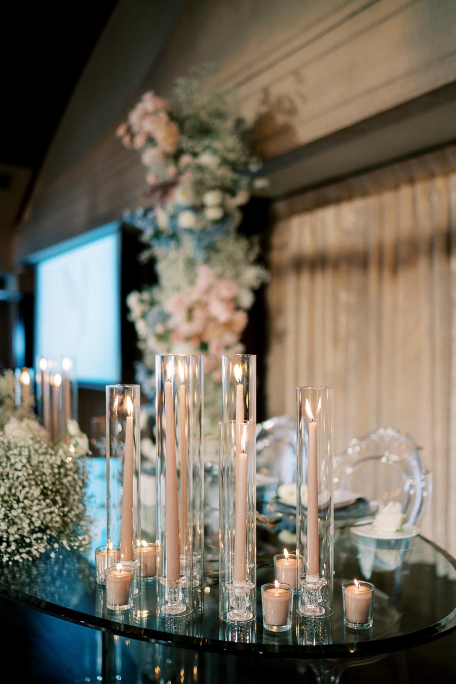 Elegant wedding reception table setting featuring tall glass candle holders with lit candles and soft floral arrangements, creating a romantic atmosphere.