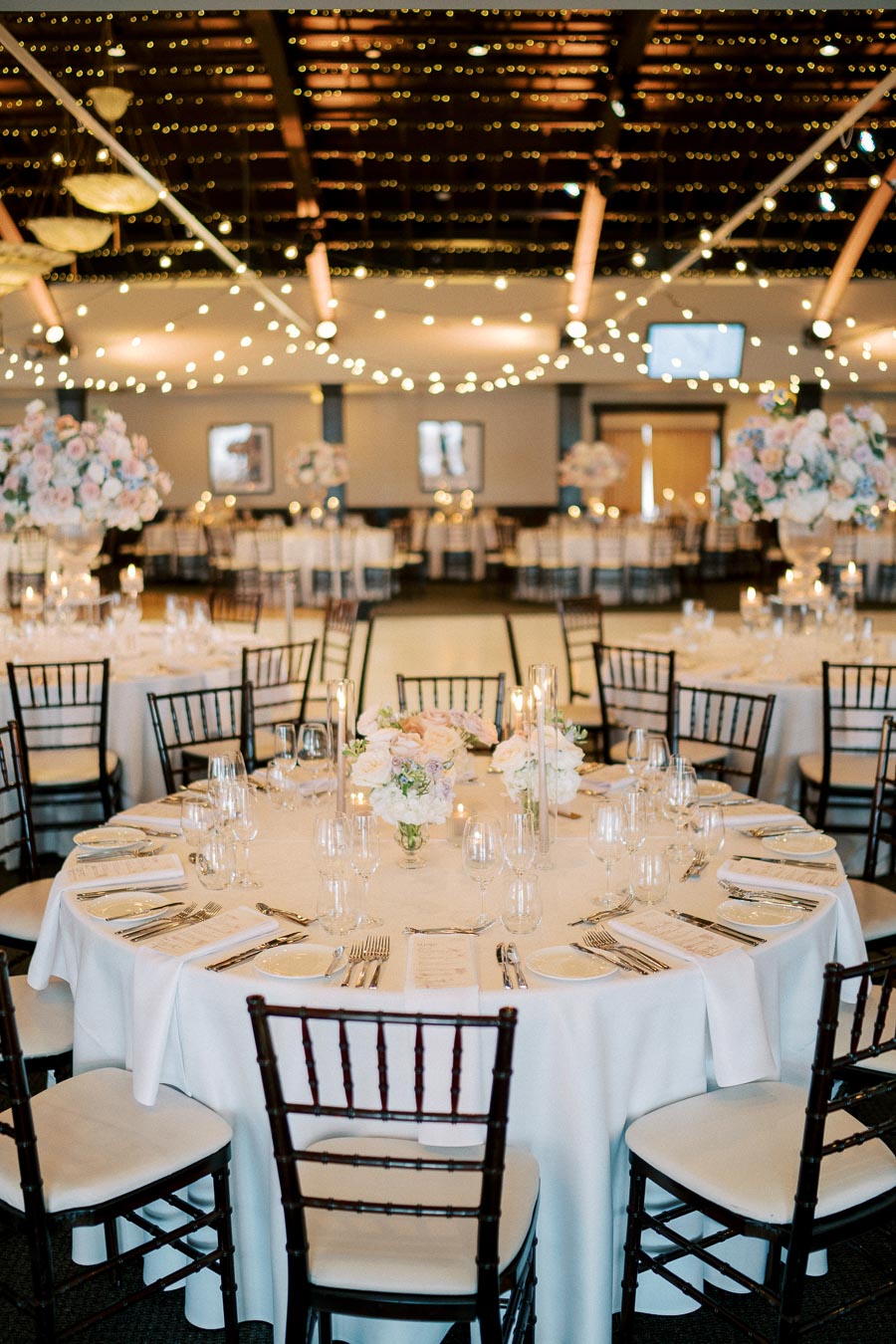 Elegant wedding reception venue with round tables set for guests, featuring white tablecloths, floral centerpieces, and ambient string lights under a vaulted wooden ceiling.