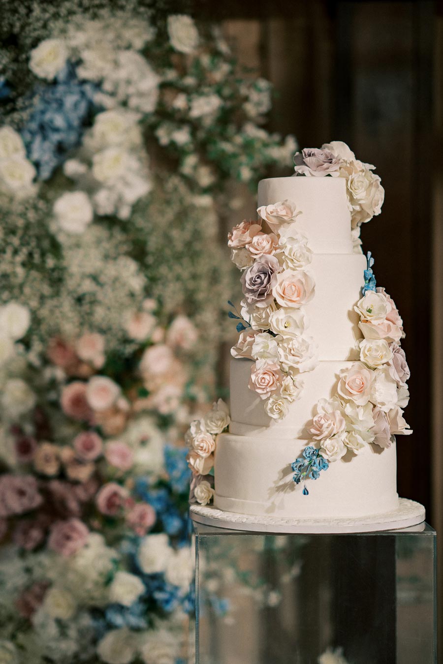 Four-tier white wedding cake adorned with cascading pastel roses and flowers, set against a blurred floral backdrop, creating an elegant celebration atmosphere.