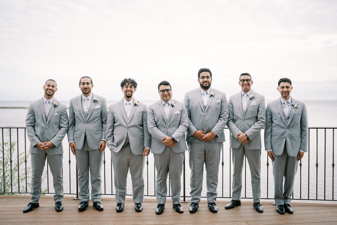 A group of seven men in matching light gray suits stand on a wooden deck by the water, posing for a formal wedding photo with a scenic waterfront background.