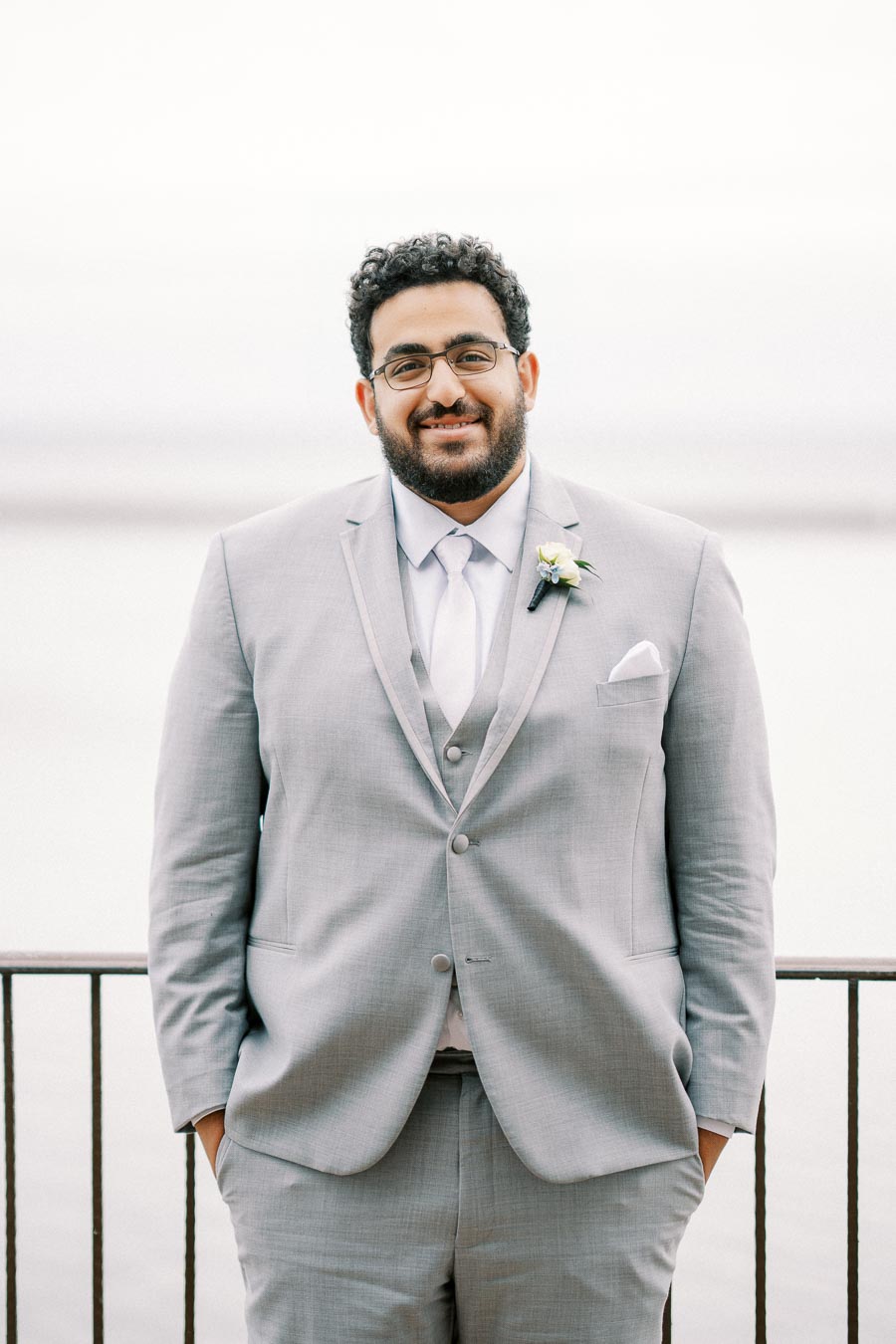 A man in a light gray suit stands confidently by a waterfront railing, wearing a boutonniere and displaying a friendly smile against a blurred background.