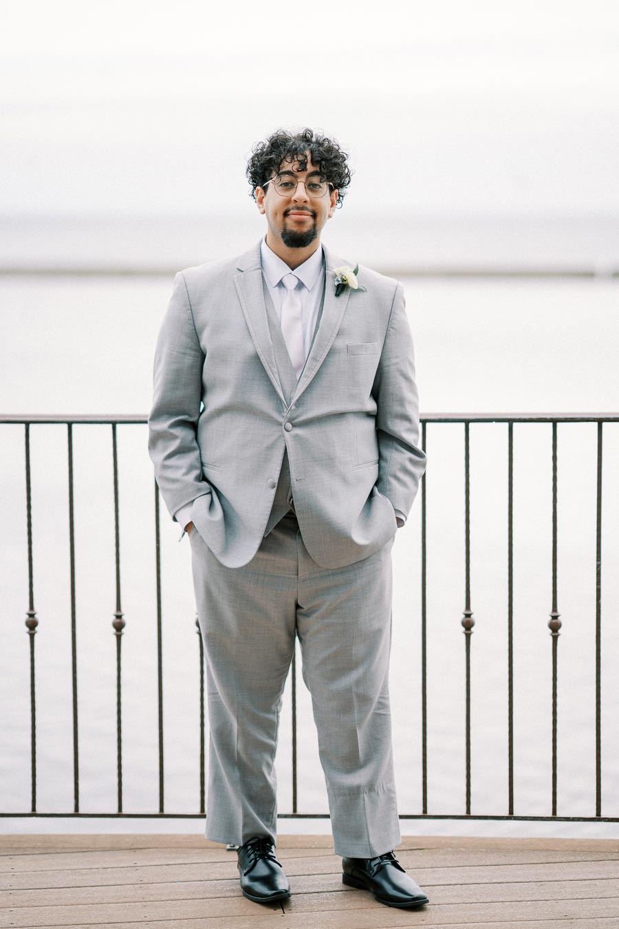 A man in a light gray suit with a white shirt and tie standing on a wooden deck in front of a railing, with a calm water backdrop.