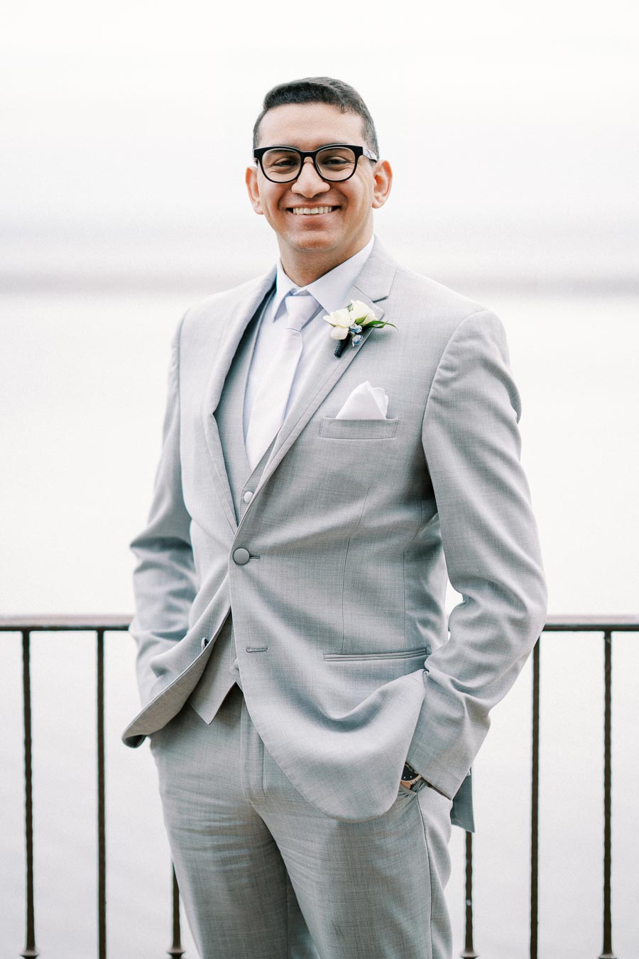 A man in a light gray suit and glasses standing by a railing, smiling, with a boutonniere on his lapel.