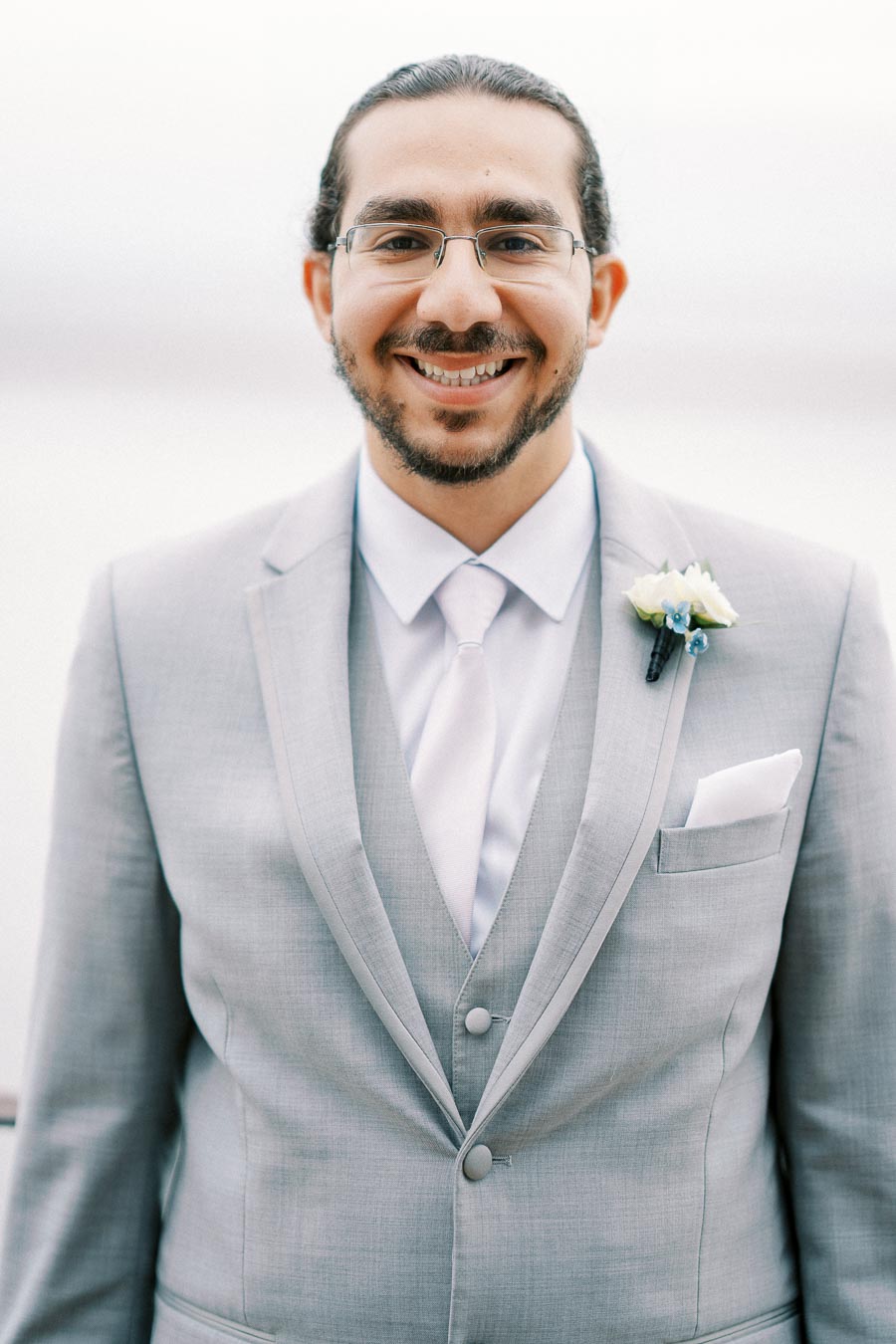 A smiling man in a light gray suit with glasses, wearing a white shirt and tie, and a boutonniere, standing against a blurred light background.