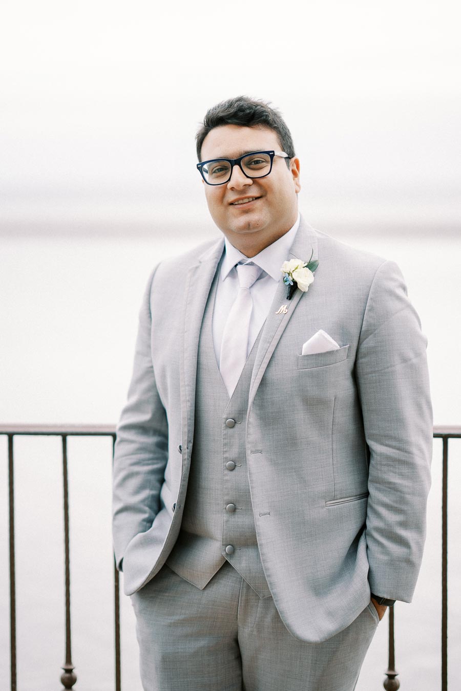 A man in a light gray suit with a white shirt and tie stands outdoors with his hands in his pockets. He is wearing glasses and smiling, with a boutonniere pinned to his lapel, against a blurred background of a railing and water.