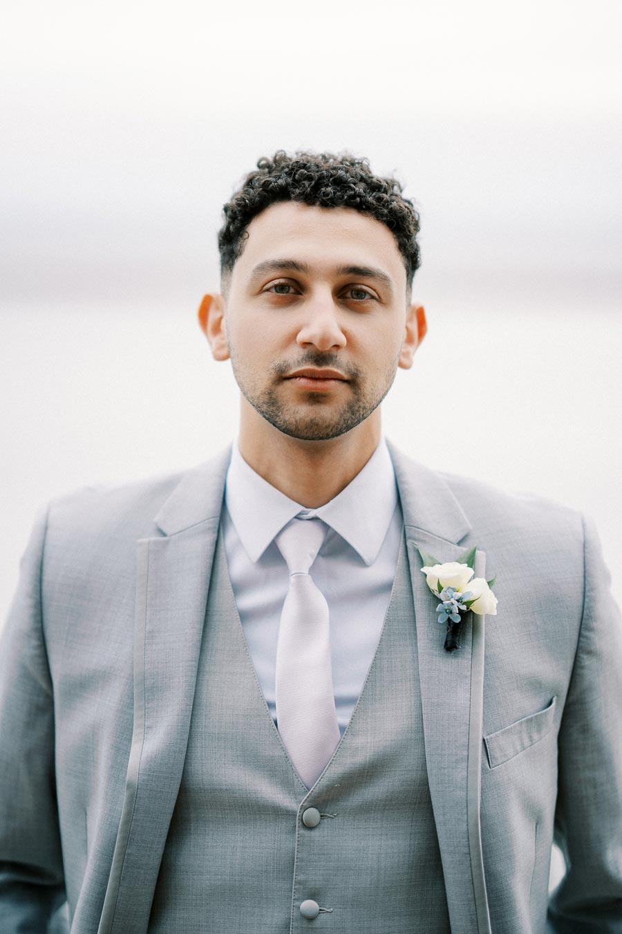 A groom in a light gray suit with a boutonniere, standing outdoors with a tranquil background.